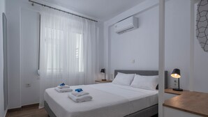 A clean, modern hotel room with a bed, white curtains, an air conditioning unit, a bedside lamp, and a wooden nightstand. - SLV Irida Apartment (Pangaio)