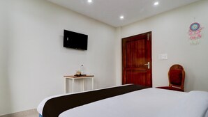 Habitación de hotel con cama, televisor montado en la pared, puerta de madera, mesita de noche con lámpara y un atrapasueños decorativo.