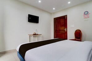 Ein Hotelzimmer mit Bett, einem an der Wand befestigten Fernseher, einer Holztür, einem kleinen Tisch mit Lampe und einem dekorativen Traumfänger.