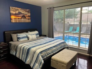 Ein Schlafzimmer mit Bett, Nachttisch, einem Fenster mit Blick auf einen Swimmingpool und einem Gemälde an der Wand.