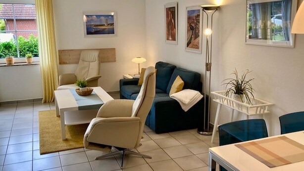 A modern living room with a sofa, armchairs, a coffee table, and a floor lamp. There are framed pictures on the wall and a window with curtains. - Ferienwohnung in Petersdorf auf Fehmarn (Fehmarn)