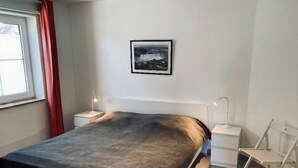 A bedroom with a bed, a nightstand, a bedside table, a chair, a window with curtains, and a framed picture on the wall. - Ferienwohnung in Petersdorf auf Fehmarn (Fehmarn)