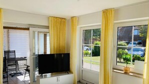A room with a TV on a cabinet, a door leading outside, and a window with curtains. - Ferienwohnung in Petersdorf auf Fehmarn (Fehmarn)