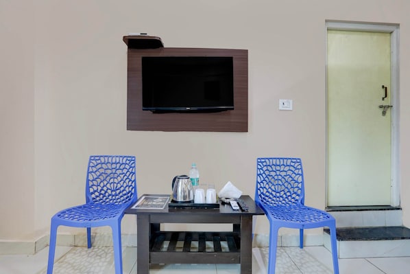 A room with a TV mounted on the wall, two blue chairs, a small table with a kettle, cups, and a remote control.