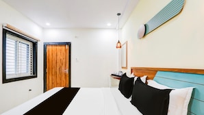 Una habitación de hotel con una cama, una ventana con persianas, una puerta de madera, una mesita de noche con lámpara y un aplique de pared.