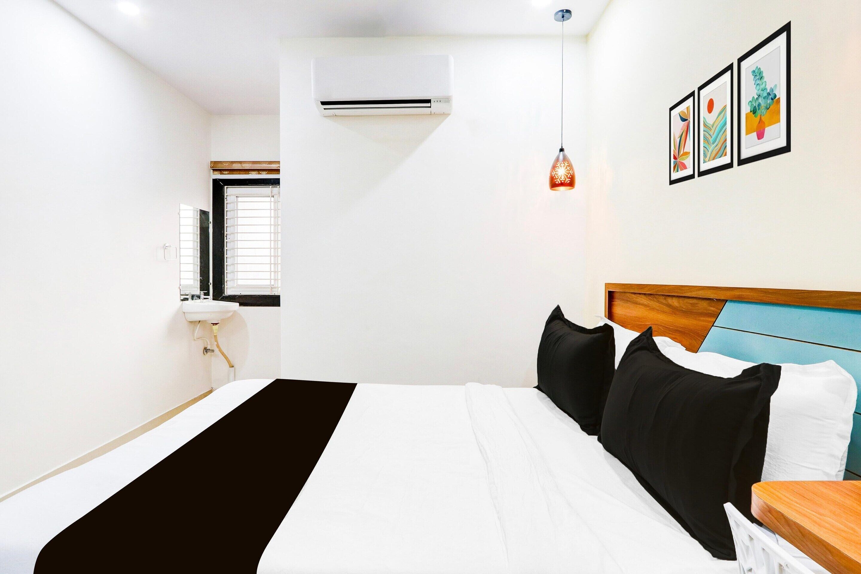 A hotel room with a bed, a bedside table, a hanging light, a window, an air conditioner, and a wall-mounted picture.