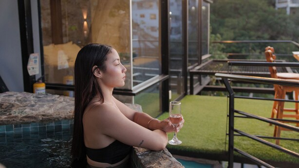 Une personne se détend au bord d’une piscine, un verre à la main, et contemple un espace verdoyant ainsi qu’un bâtiment doté de grandes fenêtres.