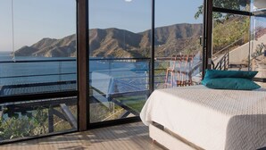 Une chambre avec une grande fenêtre donnant sur un paysage côtier, un lit recouvert d’une couette blanche et un oreiller de couleur turquoise.