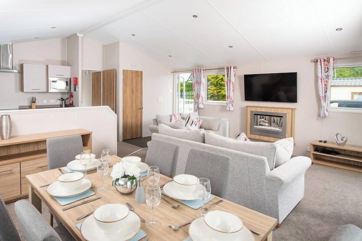 A modern kitchen with built-in appliances, a dining area with a wooden table set for four, and a living area with a sofa and TV.
