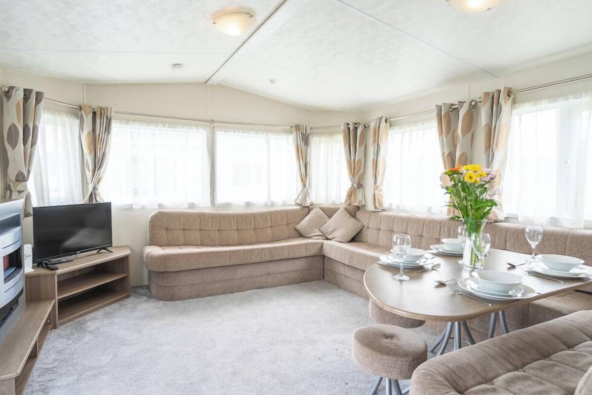 A modern living room with a sofa, a TV, a dining table set for two, and large windows with curtains.