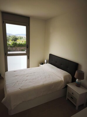 A bedroom with a bed, a nightstand, a lamp, and a view through a window. - APARTMENT WITH ROOF TERRACE L'AMPOLLA (L'Ampolla)