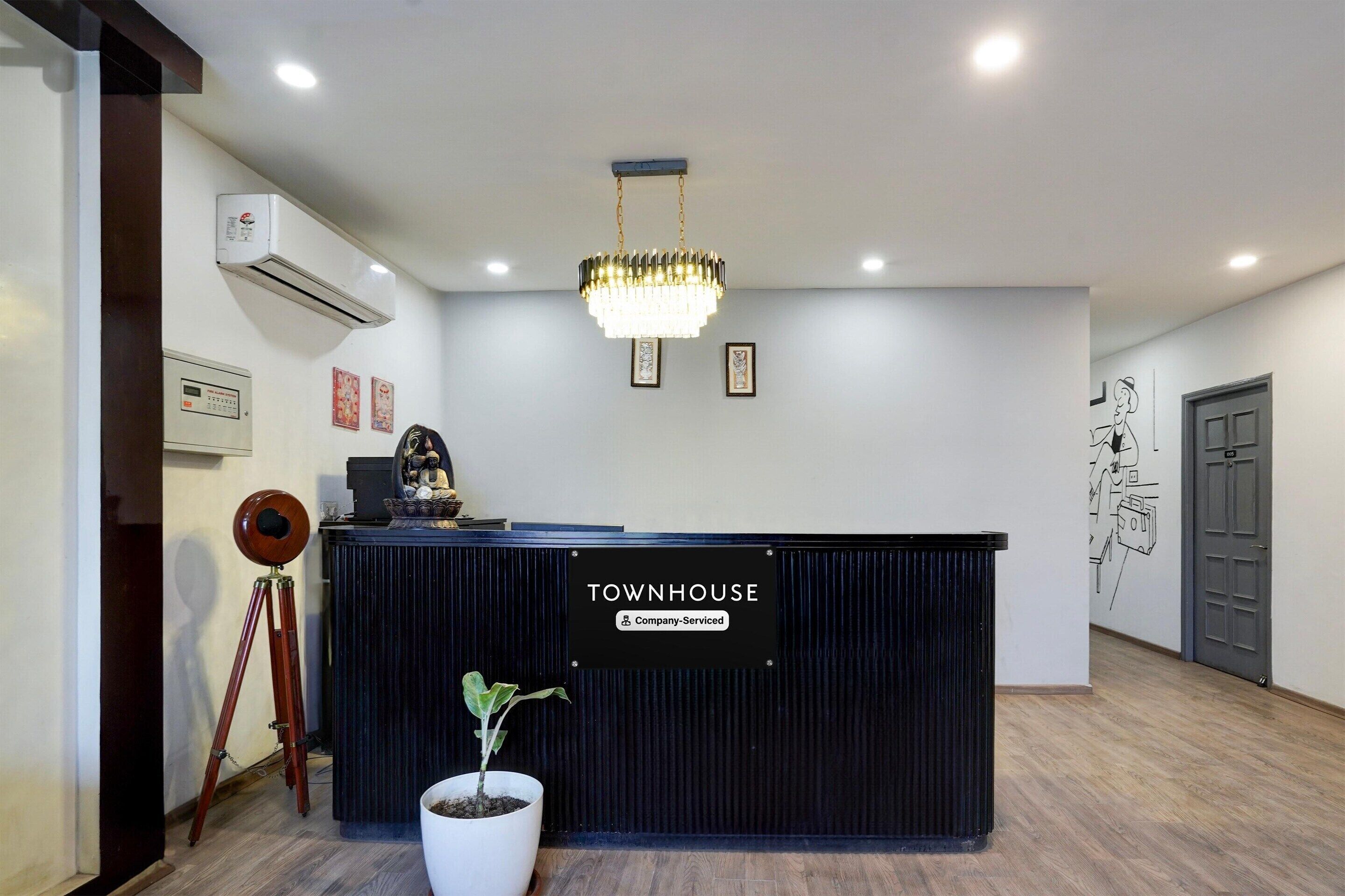 A reception desk with a sign reading 'TOWNHOUSE Company-Serviced,' a potted plant, a vintage camera on a tripod, and a wall-mounted air conditioner.
