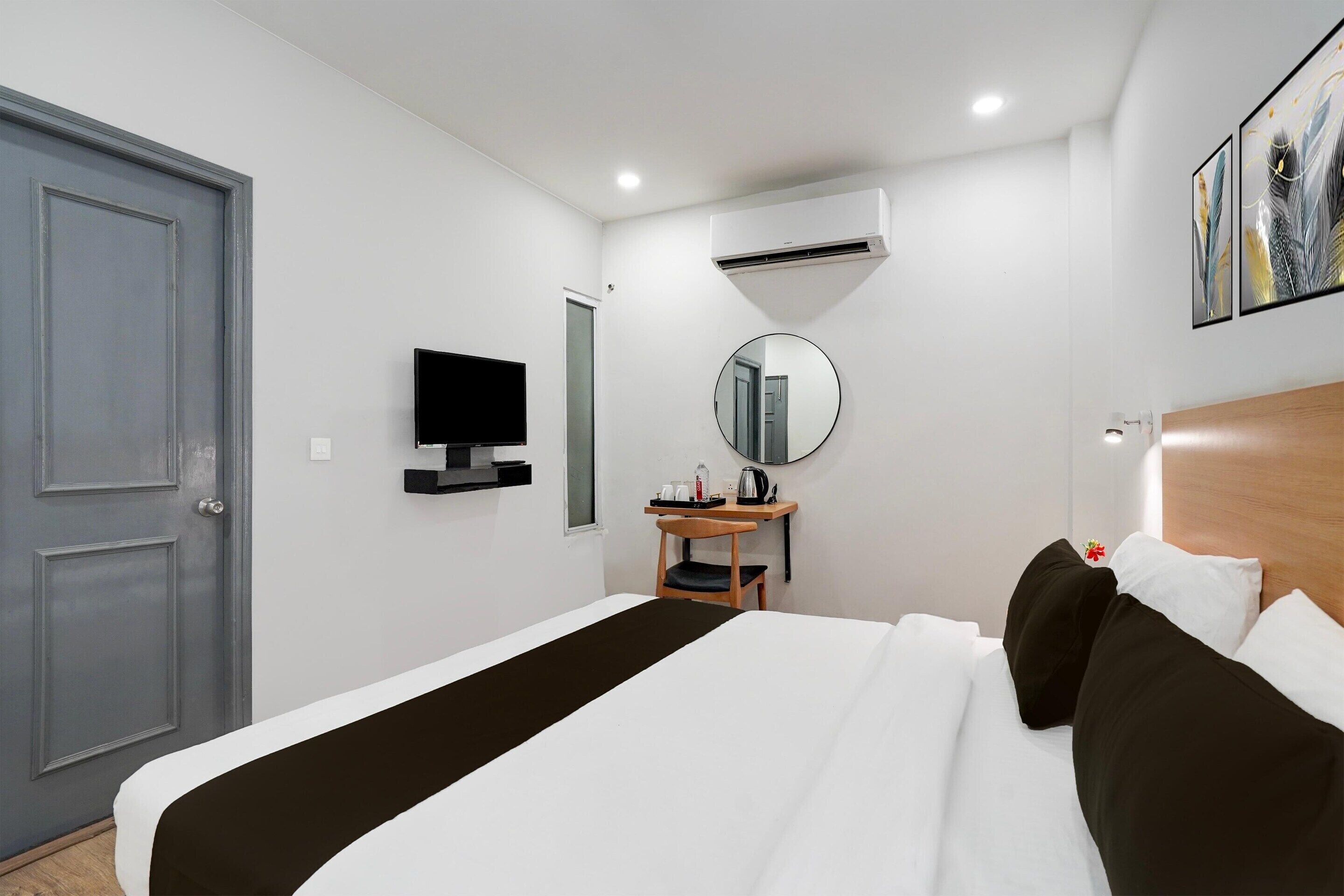 A hotel room with a bed, a desk with a chair, a television, a mirror, and an air conditioning unit.