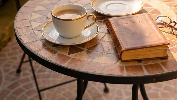 Una mesa redonda con una taza y platillo de café, un libro y un par de gafas.