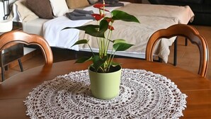 Un dormitorio con cama, una mesita de noche con una planta, una silla de madera, una mesa de comedor con un mantel de encaje y vista a una sala de estar a través de una puerta abierta.