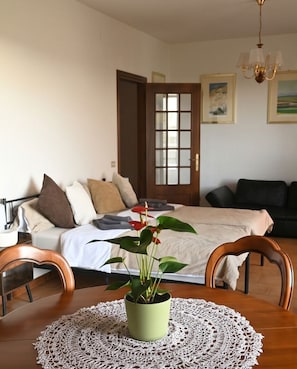 Ein Schlafzimmer mit einem Bett, einem Nachttisch mit Pflanze, einem Holzsessel, einem Esstisch mit Spitzenläufer und Blick auf ein Wohnzimmer durch eine offene Tür.