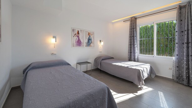 Un dormitorio con dos camas, una mesita de noche pequeña, una lámpara de pared y una ventana con cortinas.