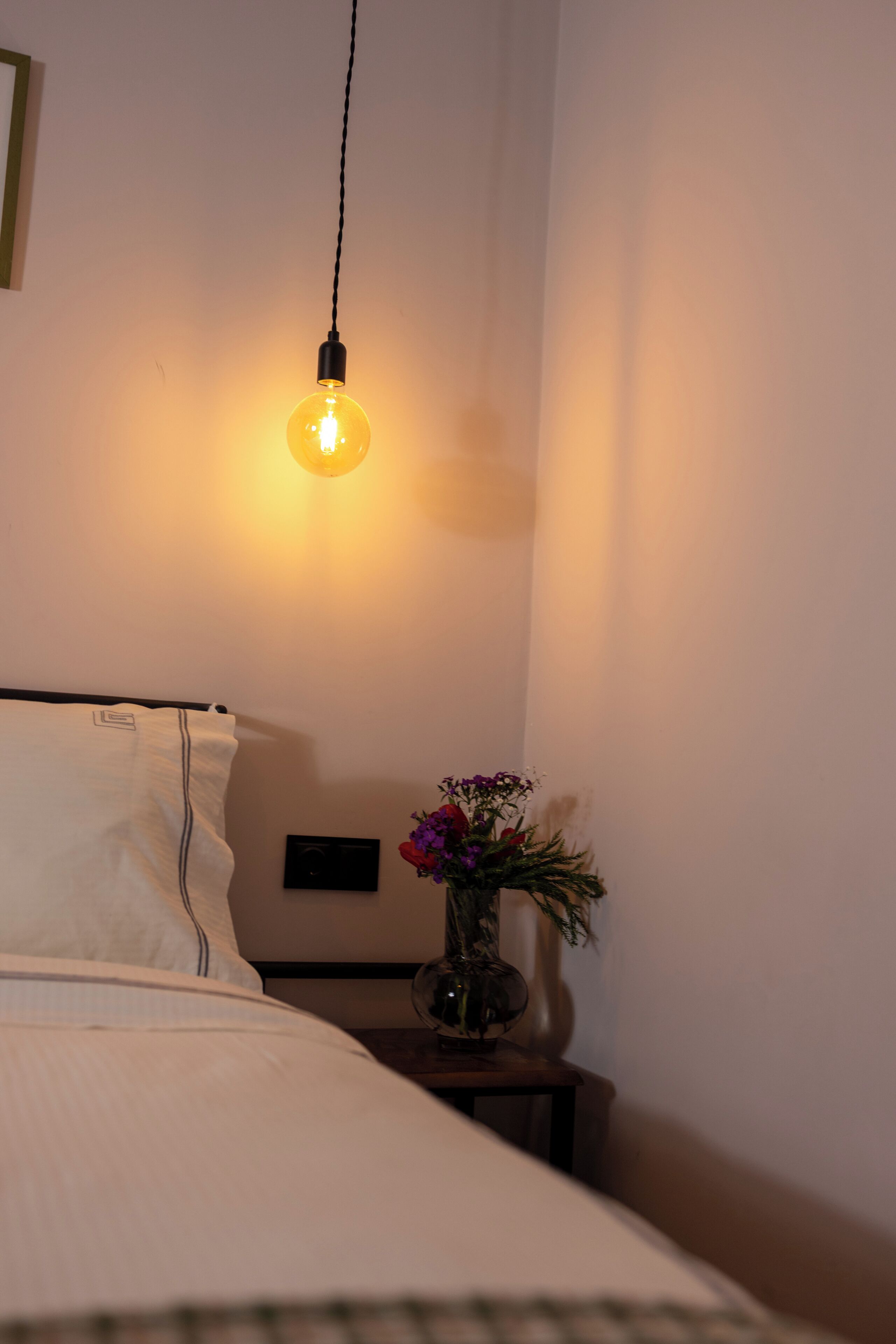A bedroom with a bed, a bedside table with a vase of flowers, and a hanging light bulb.