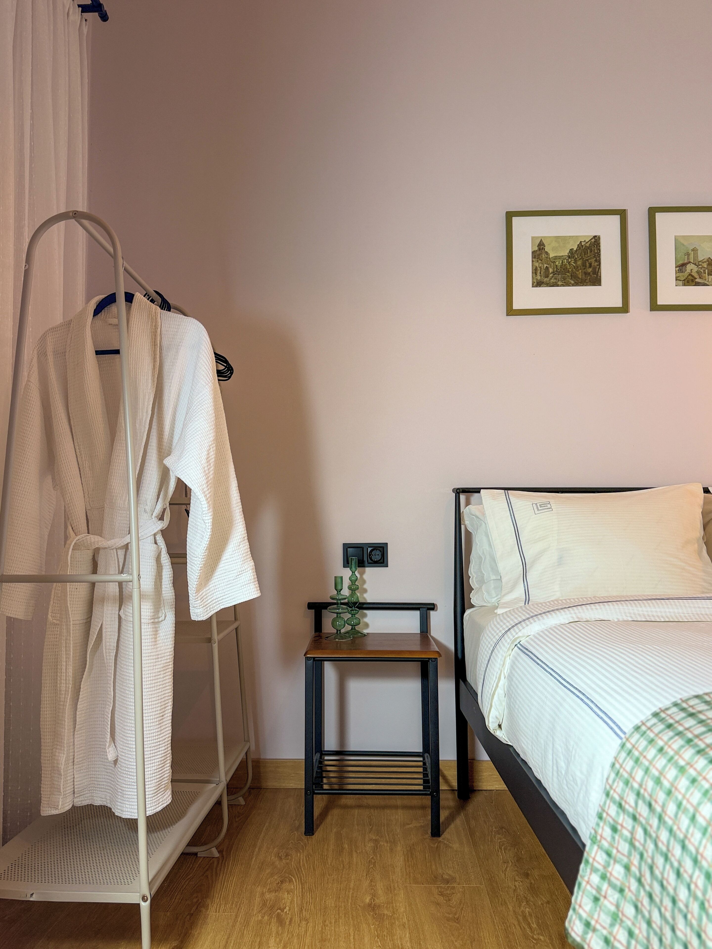 Un dormitorio con una cama, un perchero con bata, una mesita con una vela y dos cuadros en la pared.