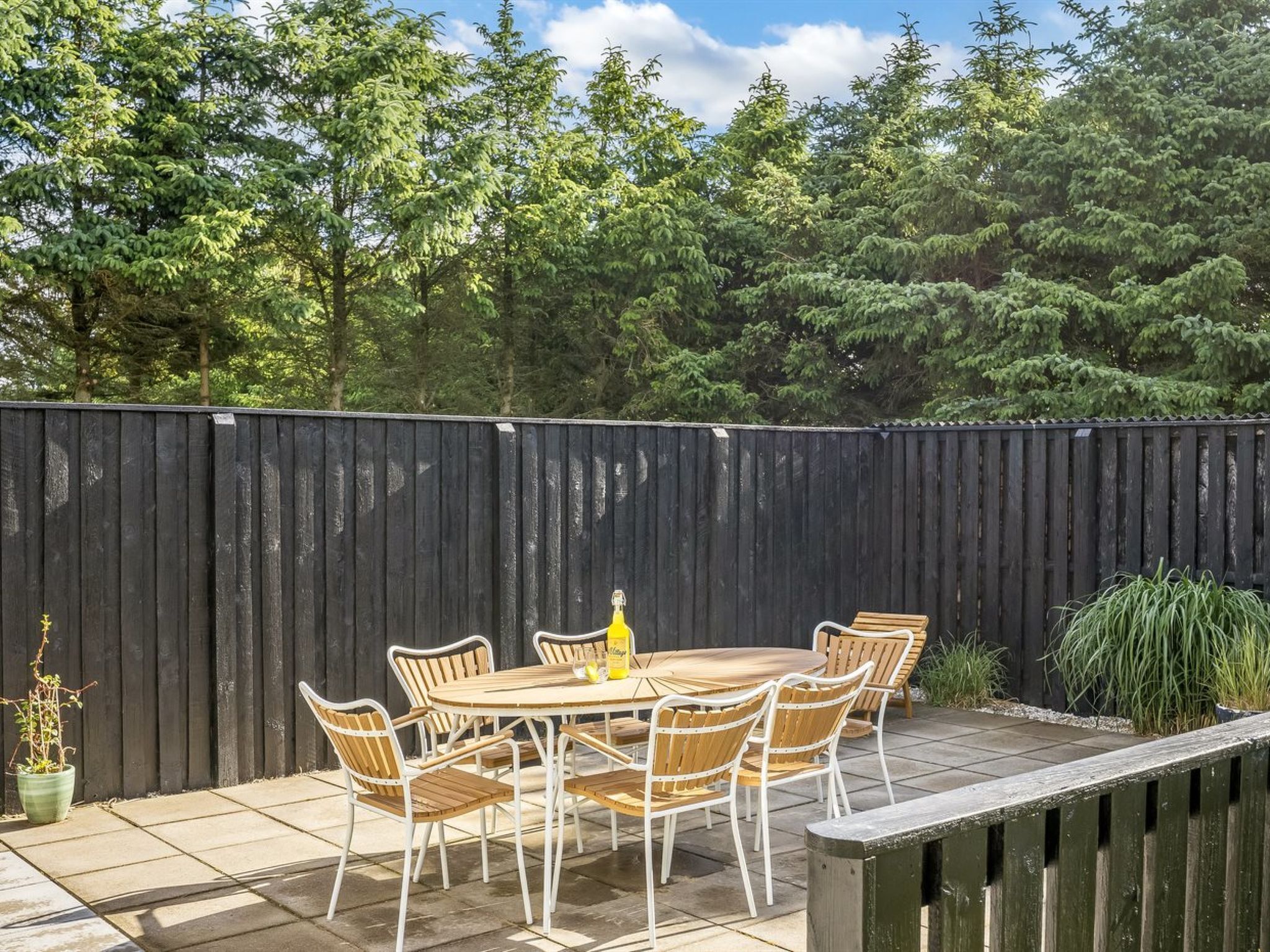 Een terras buiten met een ronde tafel en stoelen, omringd door een houten hek en groen.