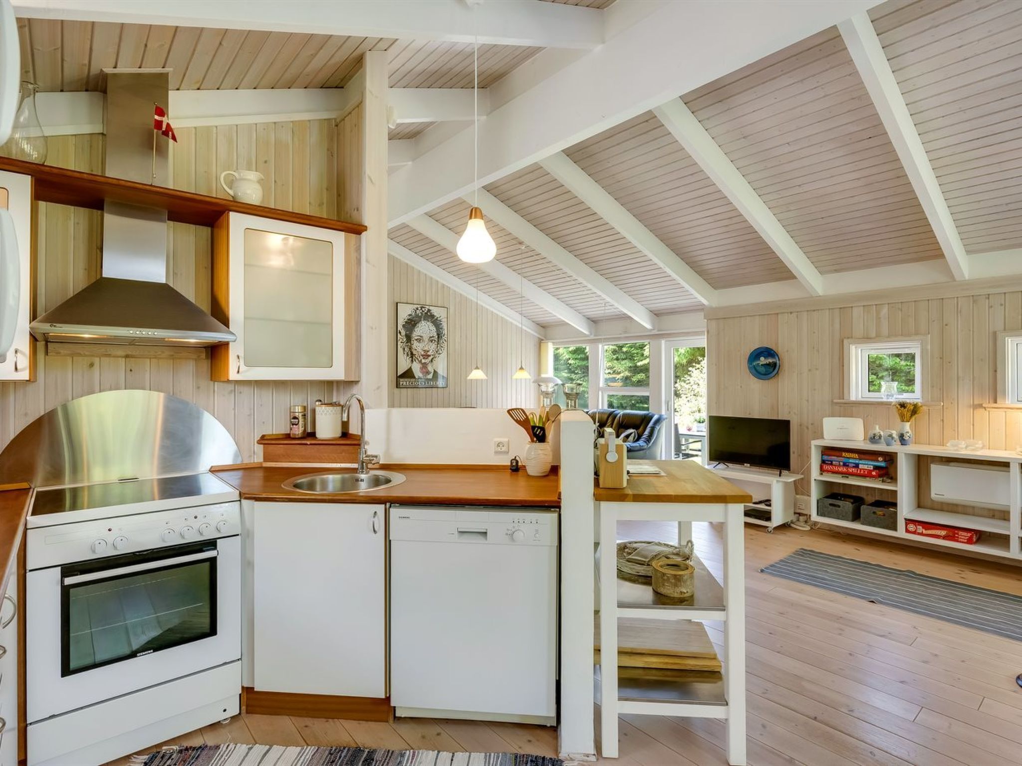 Une cuisine compacte équipée d’une cuisinière, d’un four et d’un lave-vaisselle, avec un plancher en bois et un coin repas comprenant une télévision et des livres.