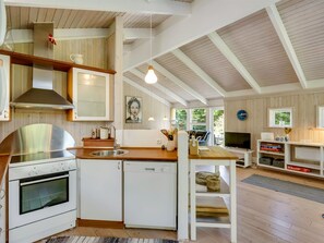Cocina compacta con estufa, horno y lavavajillas, piso de madera y comedor con televisor y libros.