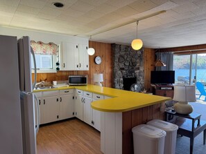 A kitchen with white cabinets, a yellow countertop, a stone fireplace, a television, and a view of the outdoors. - Happiness Awaits on Lake Winnipesaukee-By Break Away (Meredith)