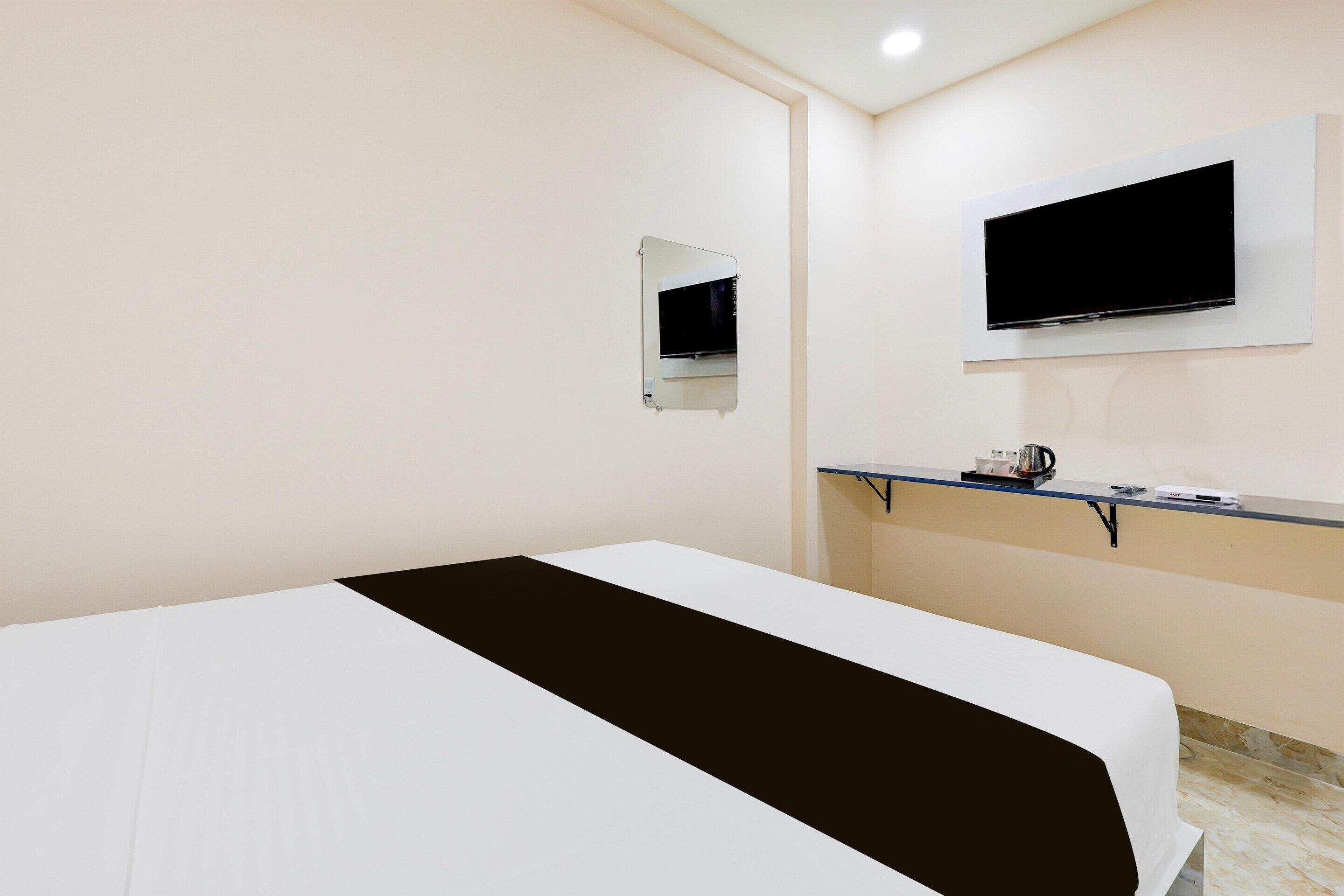 A hotel room with a bed, a TV mounted on the wall, a small mirror, and a shelf with a kettle and other items.