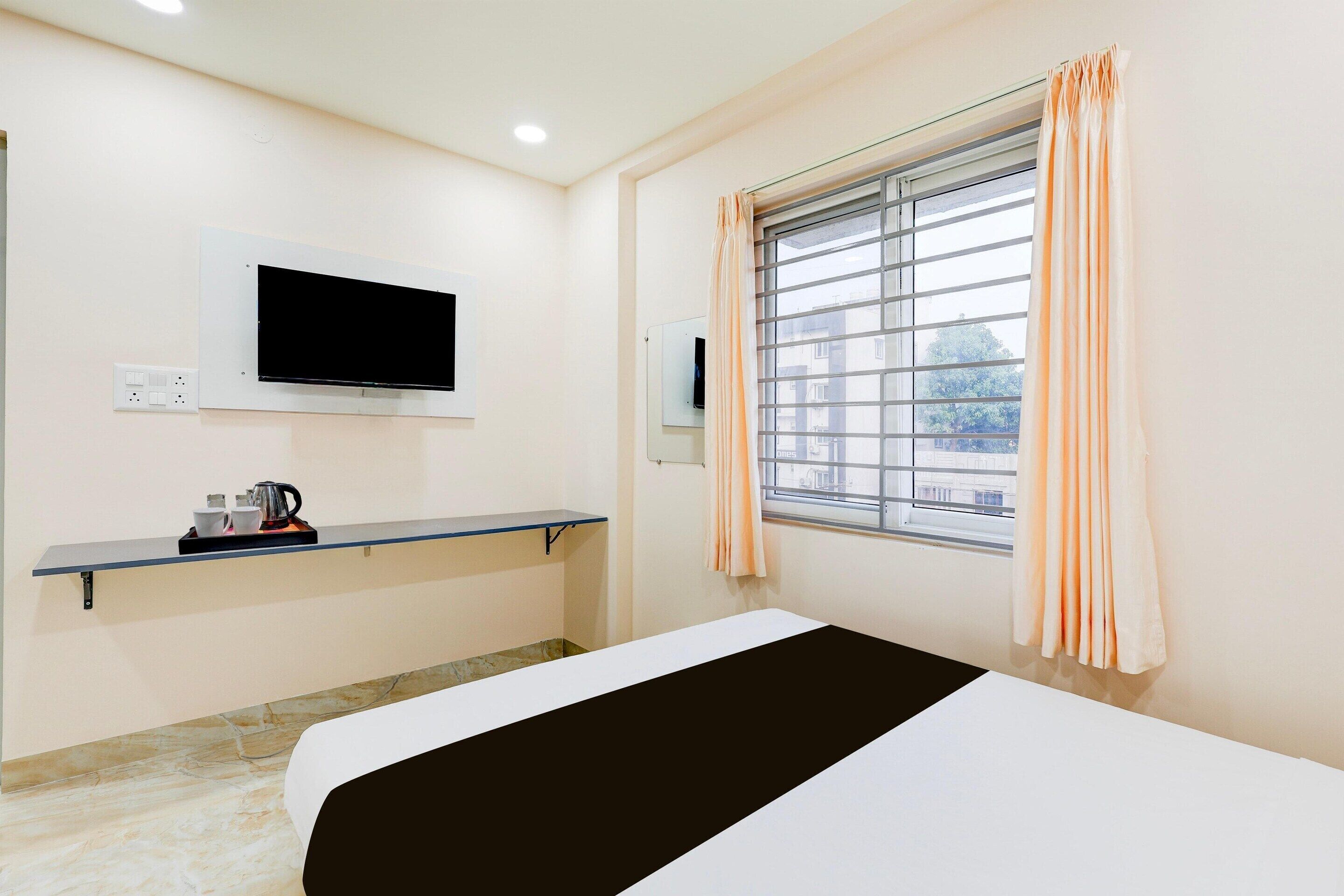 A hotel room with a bed, a mounted TV, a window with curtains, a shelf with a kettle, and a wall-mounted power outlet.