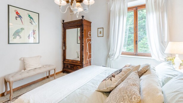 A bedroom with a wooden ceiling, a large bed with patterned bedding, a bench, a mirror, and a window with curtains. - Apartment in Vorno Near Lucca City Center (Capannori)