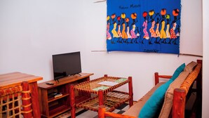 Una habitación con televisión, una silla de mimbre, un sofá con cojines y un tapiz en la pared.