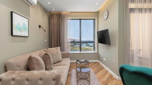 A living room with a sofa, a coffee table, a TV, and a large window with a view. - XIO Apartments | Stylish Comfort with Balconies & Full Kitchens (Bar)
