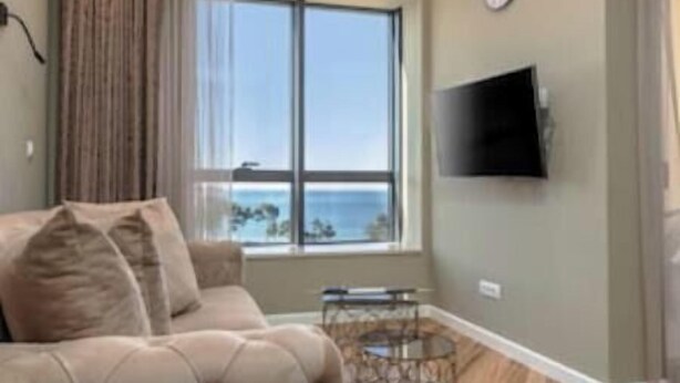 A living room with a sofa, a TV, a window with a view, and a decorative metal stand. - XIO Apartments | Stylish Comfort with Balconies & Full Kitchens (Bar)