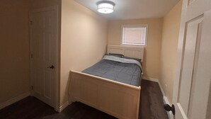 A small bedroom with a single bed, a door, a window with blinds, and a ceiling light fixture.