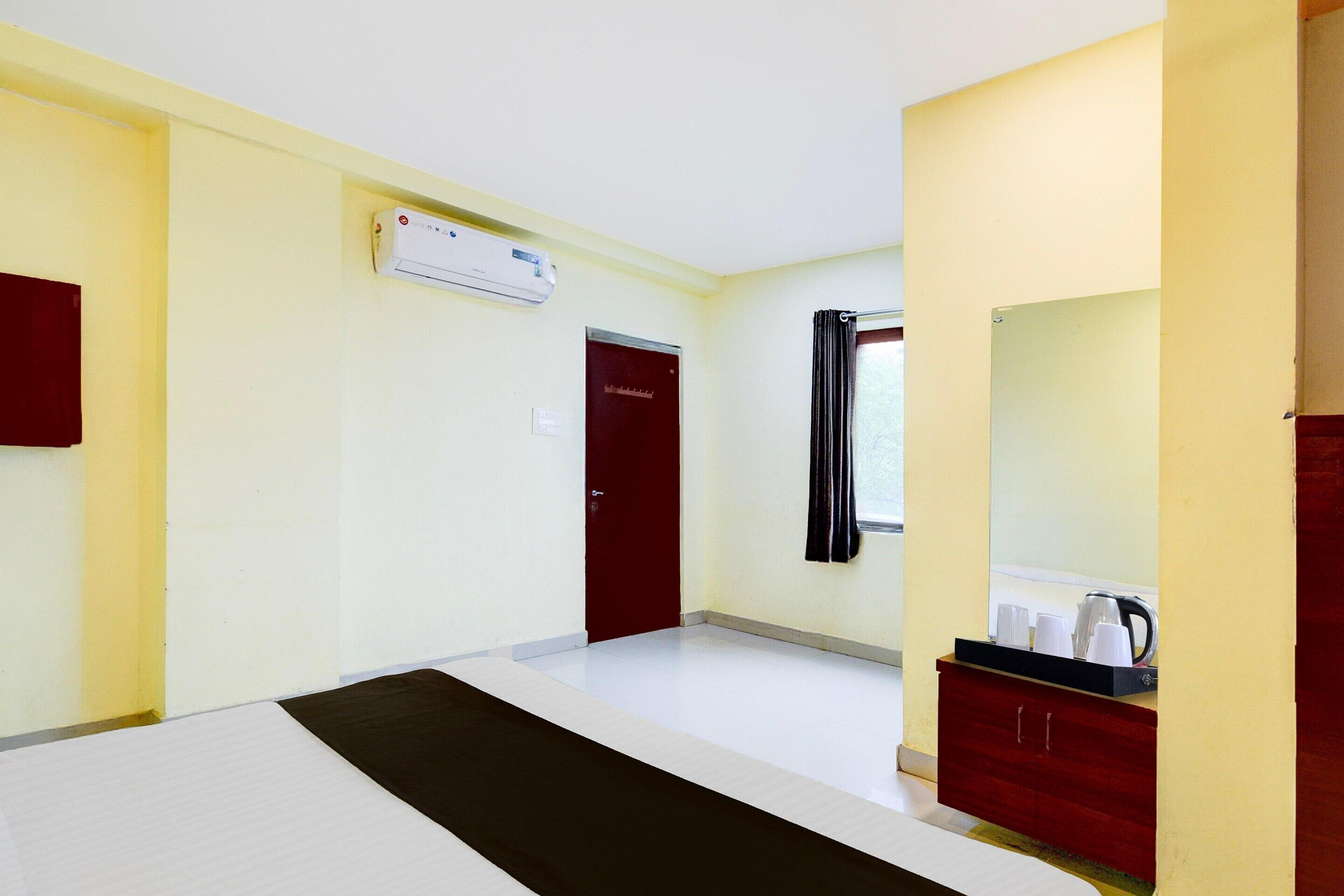 Une chambre d’hôtel avec un lit, une porte rouge, un climatiseur blanc, une fenêtre avec des rideaux et une commode en bois sur laquelle se trouvent une bouilloire et des tasses.
