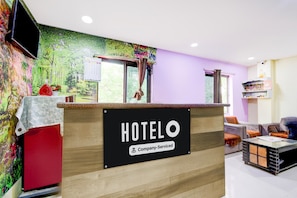 Un mostrador de recepción de hotel con un cartel que dice 'HOTELO Company-Serviced', un televisor, una ventana con cortinas y una zona de estar con un sofá y una mesa.