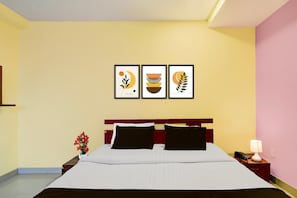 Quarto de hotel com uma cama, uma mesa de cabeceira com abajur, um vaso com flores e três quadros na parede.