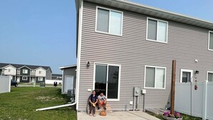 A two-story residential building with a front porch, a garage, and a small front yard with a wooden planter.