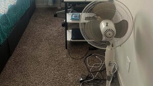 A bedroom with a bed, a desk with a computer, a fan, and a painting on the wall.