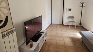 Un soggiorno con televisore a schermo piatto su un supporto bianco, un attaccapanni, una poltrona e un letto.