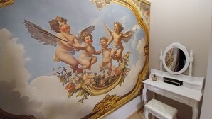 Een kamer met een grote muurschildering van cherubijnen op het plafond, een witte wastafel met spiegel en een klein bankje.