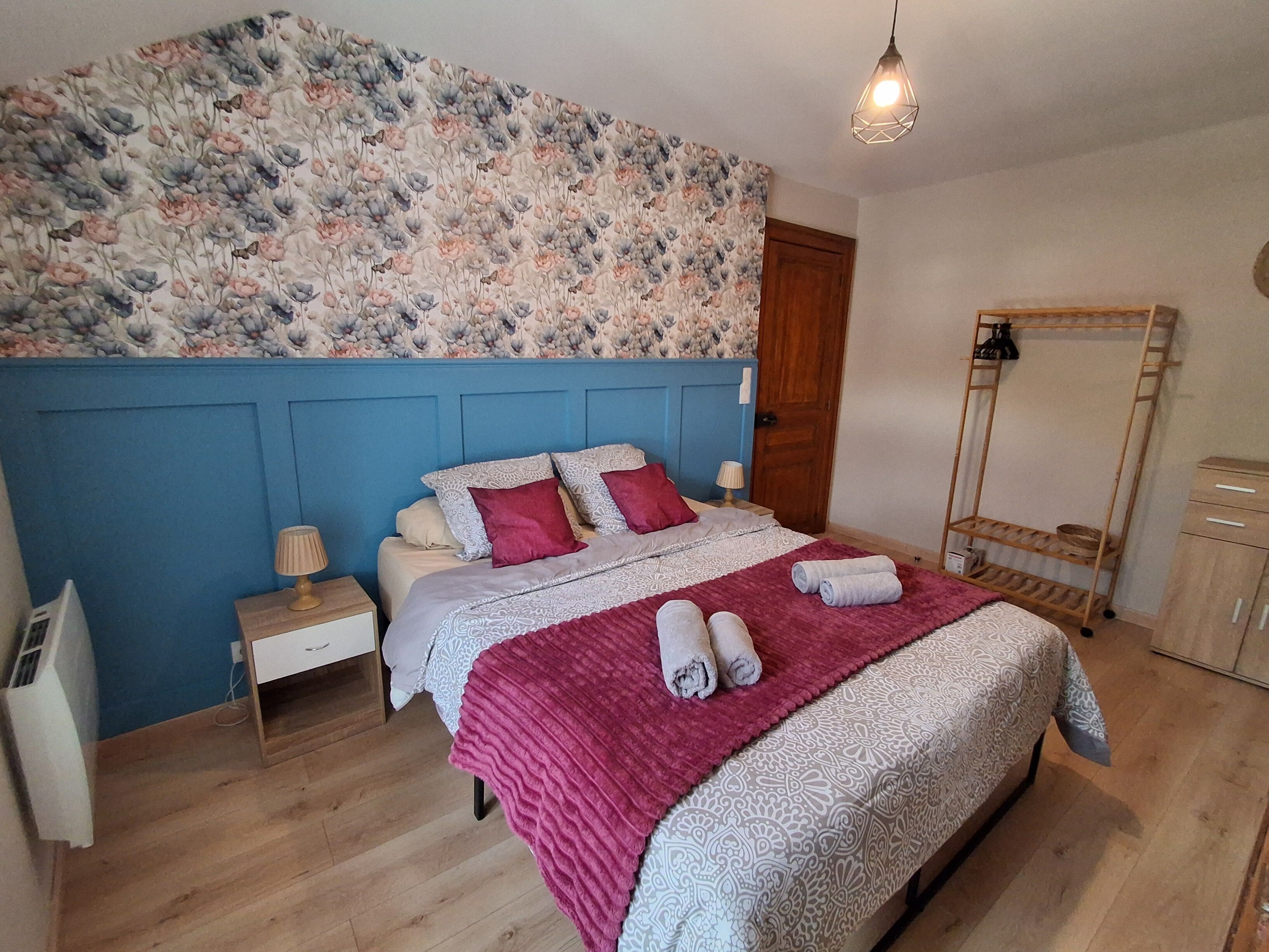 A bedroom with a blue headboard, a bed with patterned bedding, a wooden floor, a nightstand, a lamp, a mirror, and a wooden shelf.