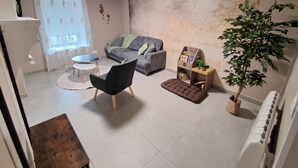 Een woonkamer met een grijze bank, een zwarte fauteuil, een klein rond tafeltje, een bruin poefje, een houten bijzettafeltje met boeken, een potplant en een radiator.