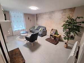 A living room with a grey sofa, a black armchair, a small round table, a brown ottoman, a wooden side table with books, a potted plant, and a radiator. - Un Air de Poudlard (Bogny-sur-Meuse)