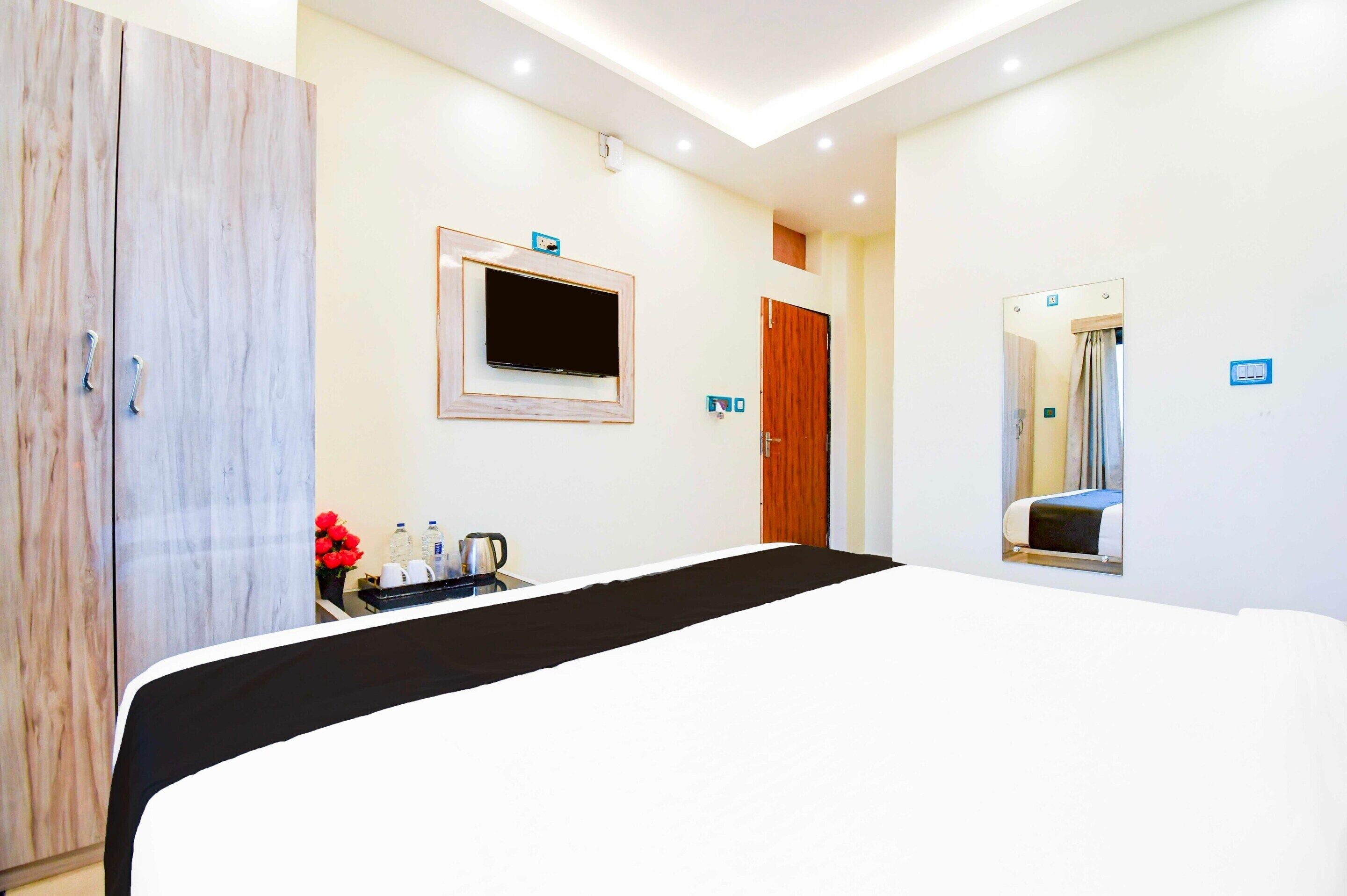 Habitación de hotel con cama, televisor, armario, espejo y una mesita con hervidor y flores.