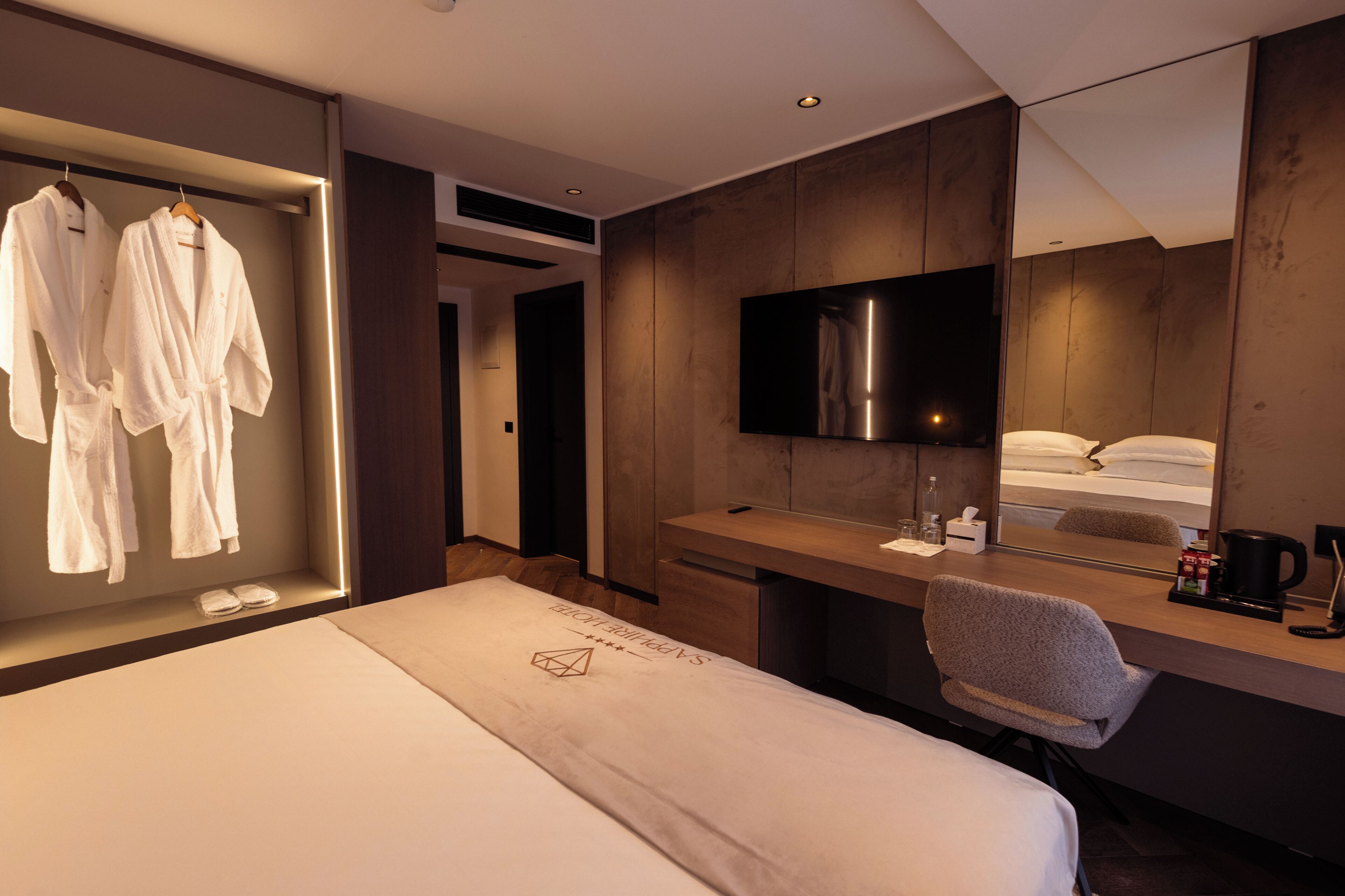 A hotel room with a bed, a desk with a mirror, a TV, and a seating area.