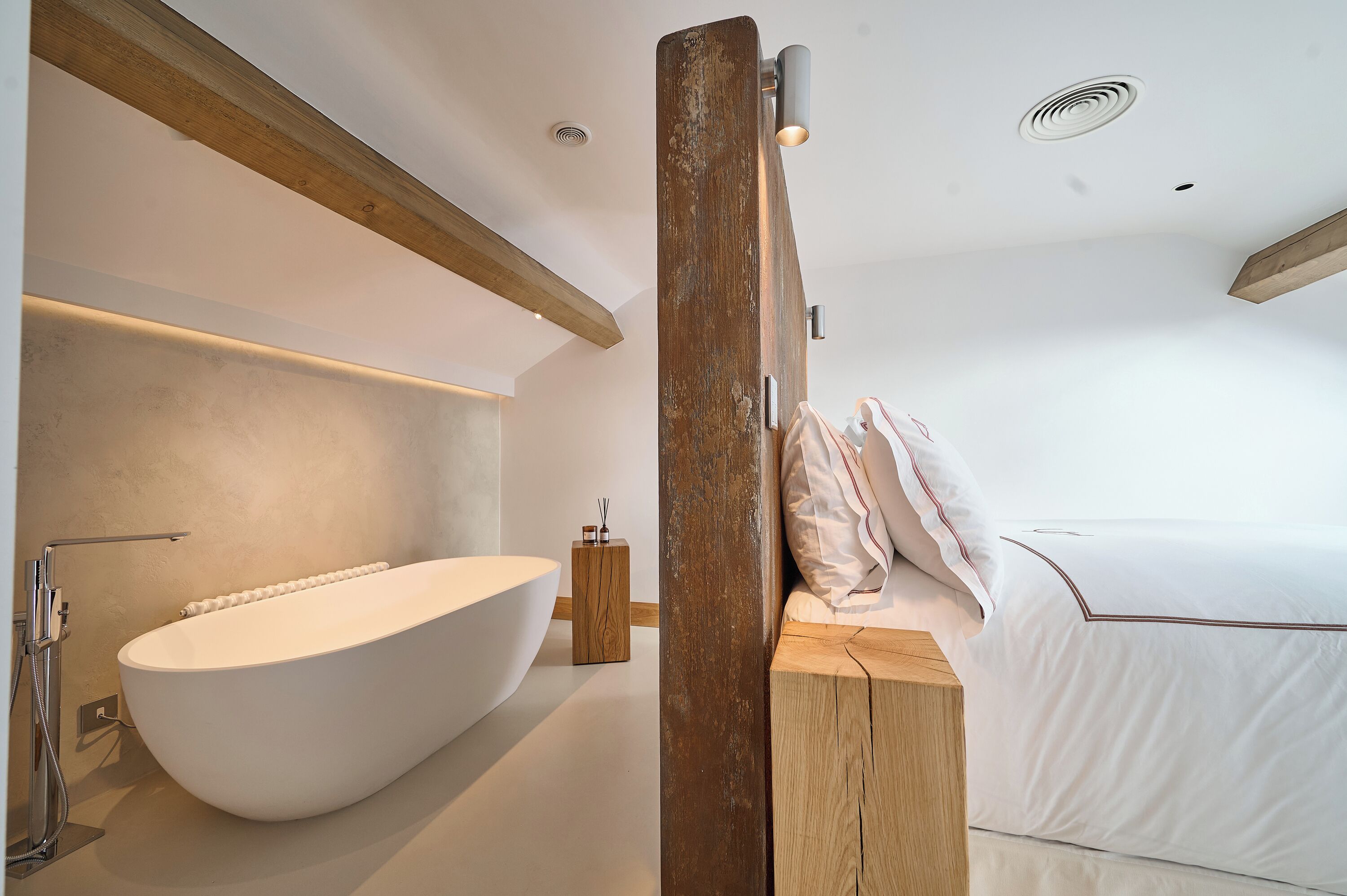 Un baño moderno con una bañera independiente, una mesita de noche de madera y una cama con ropa de cama blanca.