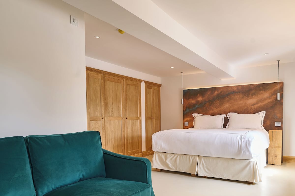 Una habitación de hotel moderna con un sofá verde azulado, una mesa de centro de madera con un florero, una cama grande con ropa de cama blanca y un cabecero de madera.