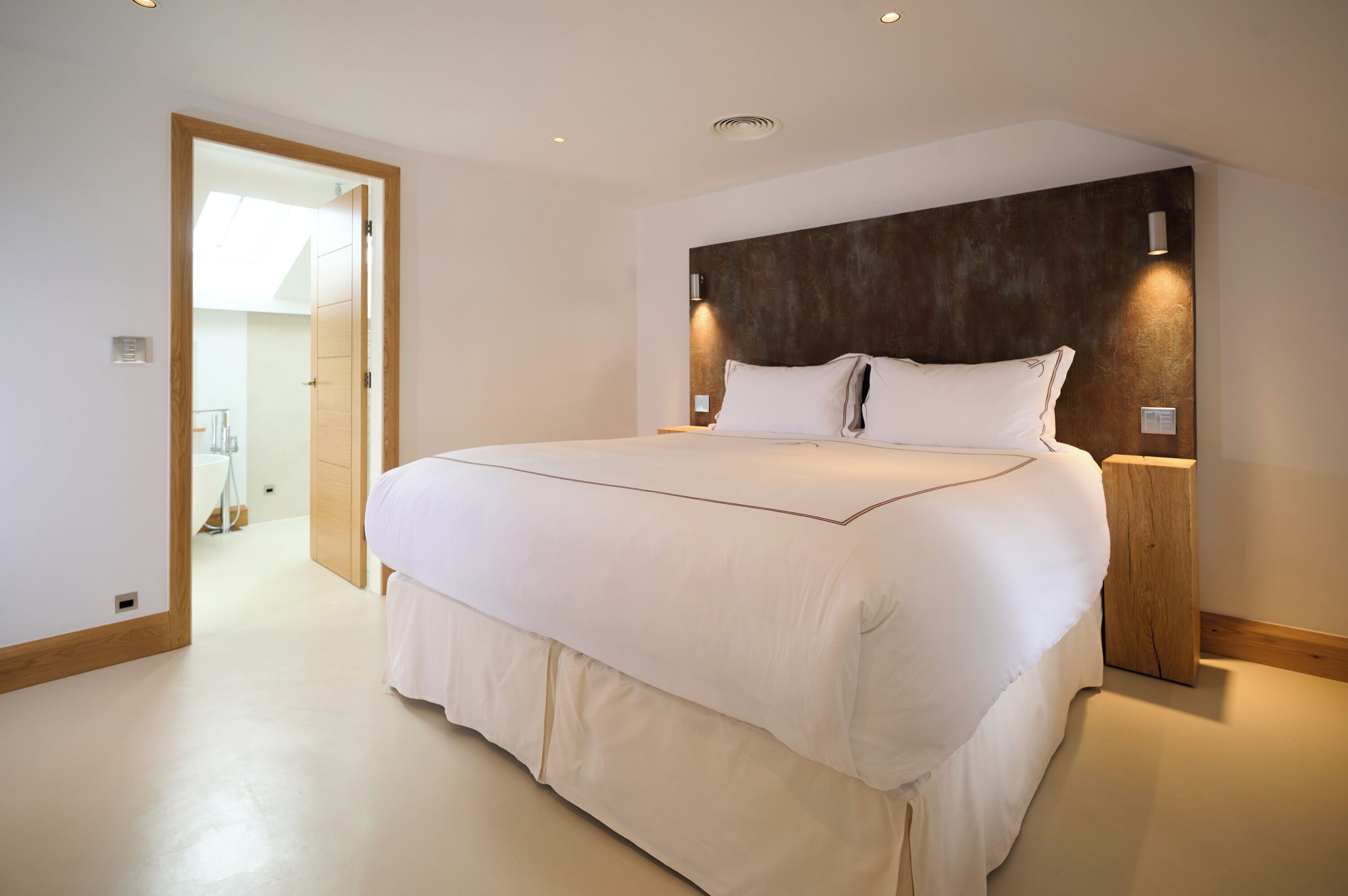 Uma cama grande com cabeceira de madeira, luminárias laterais e um banheiro visível ao fundo.
