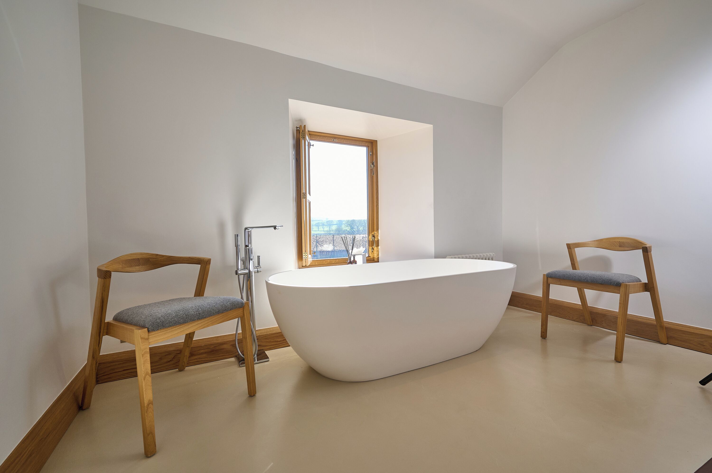 Un baño moderno con una bañera independiente, dos sillas de madera, un ventanal con vistas y un lavamanos blanco.